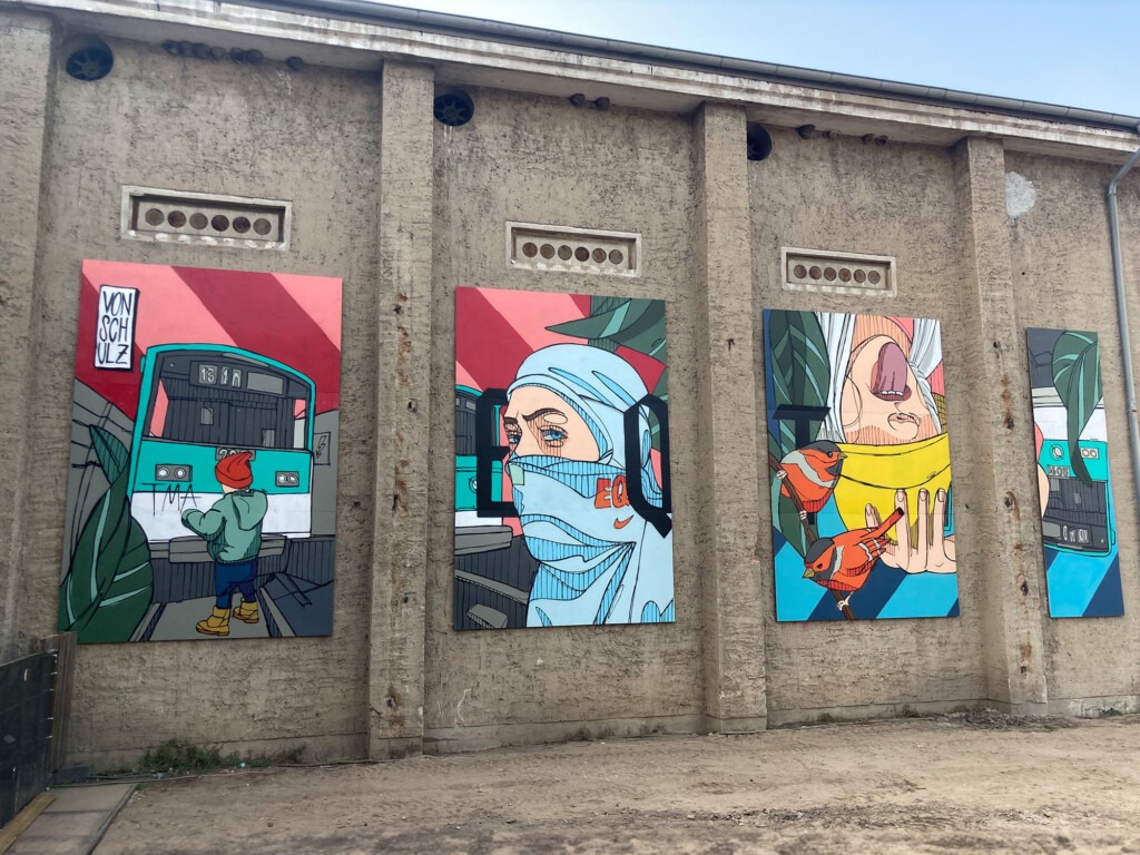 Vier aufeinander abgestimmte Wandpaneele, je 4 Meter hoch, mit urbanen Motiven, darunter Menschen und Vögel, auf dem Gelände des Splash Festivals.