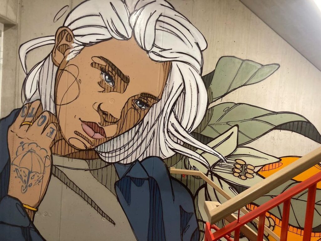 Wandgemälde einer Frau mit weißen Haaren und einem farbenfrohen Vogel auf einer Treppe, umgeben von üppigen Pflanzen.