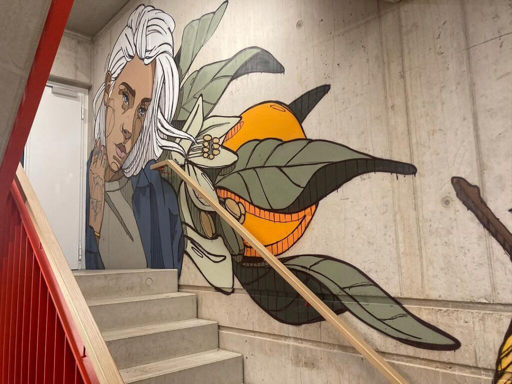 Wandgemälde einer Frau mit weißen Haaren und einem farbenfrohen Vogel auf einer Treppe, umgeben von üppigen Pflanzen.