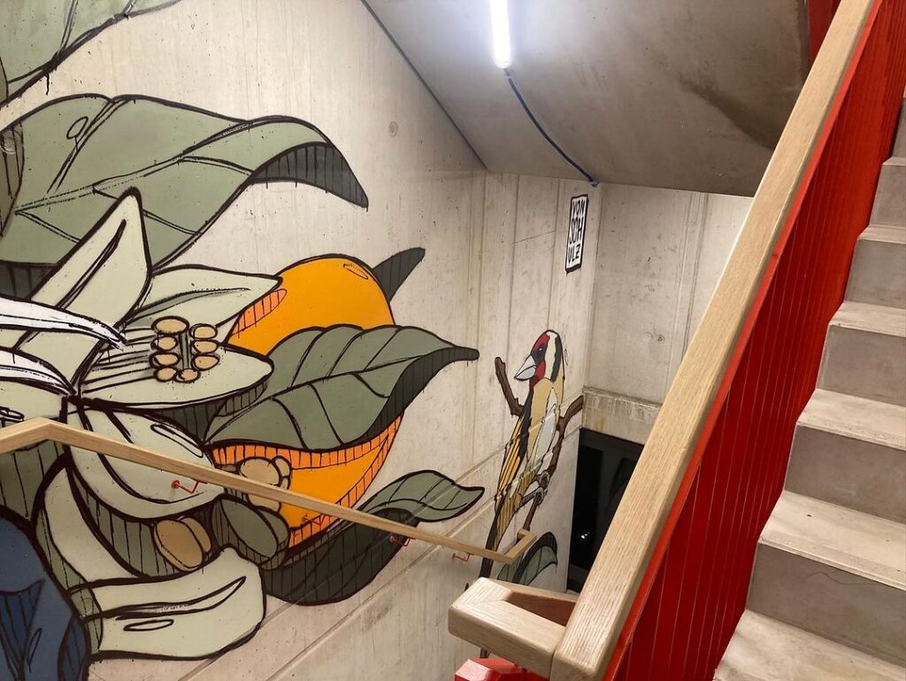 Wandgemälde einer Frau mit weißen Haaren und einem farbenfrohen Vogel auf einer Treppe, umgeben von üppigen Pflanzen.
