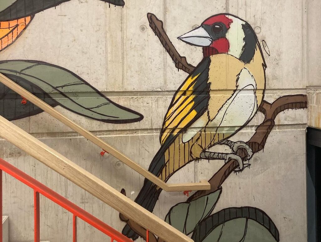 Wandgemälde einer Frau mit weißen Haaren und einem farbenfrohen Vogel auf einer Treppe, umgeben von üppigen Pflanzen.