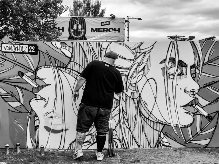 Street-Art-Künstler in Aktion, gestaltet ein schwarz-weißes Wandgemälde mit abstraktem, geometrischem Design und stilisiertem weiblichen Gesicht bei einem Kultur- oder Musikfestival, umgeben von Spraydosen auf dem Boden.